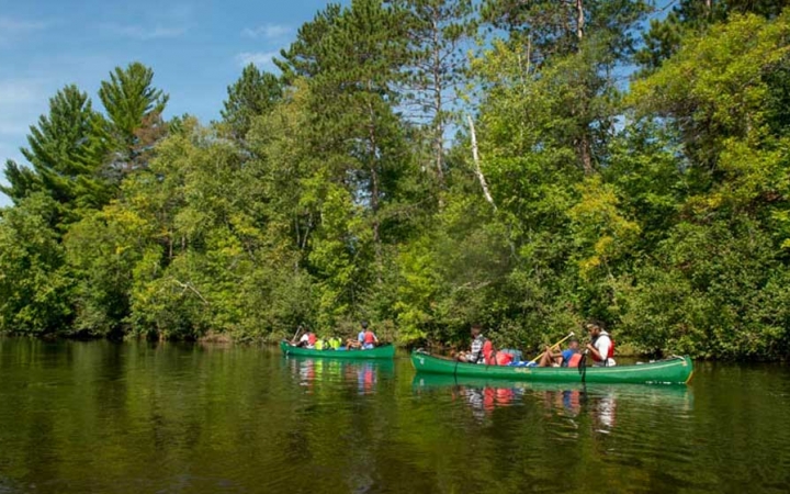 People paddle two canoes on calm water near a tree-lined shore.